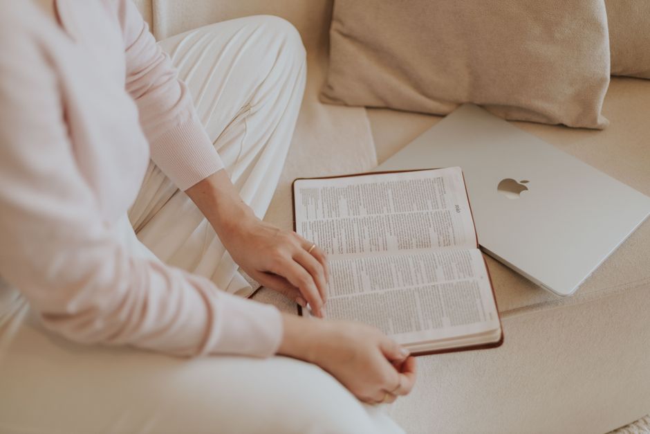 A woman in casual attire reads a book beside a laptop while sitting on a cozy sofa.