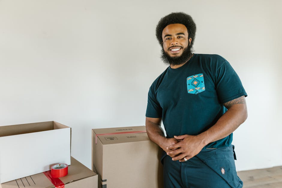 A cheerful man with afro hair standing next to moving boxes indoors, smiling.