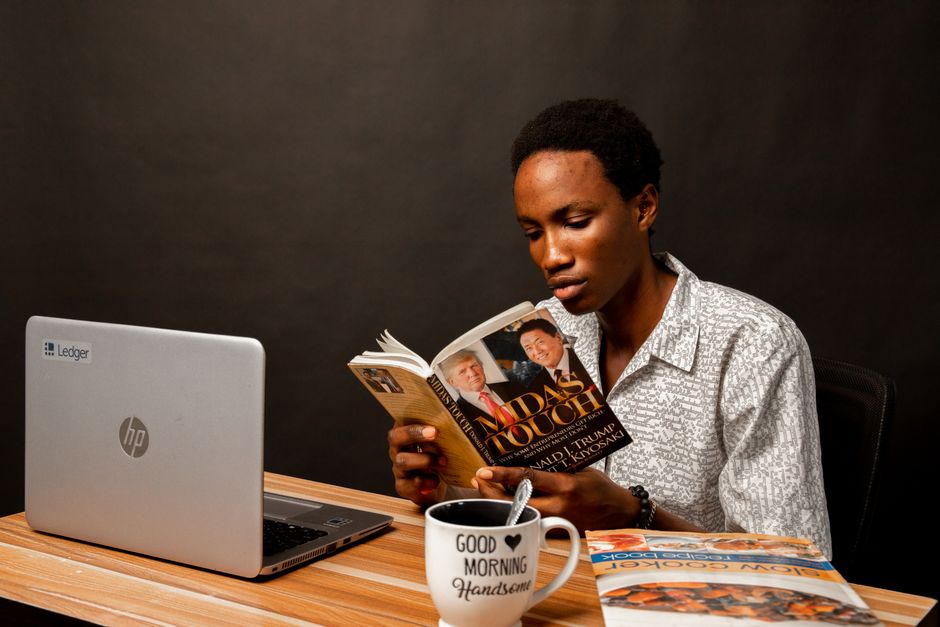 Young man reading a book at his desk with a laptop and coffee mug.