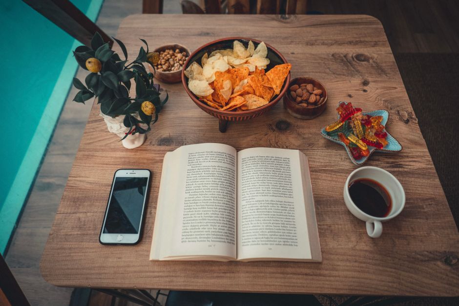 Top view of a wooden table with a book, snacks, coffee, and smartphone, creating a cozy reading setup.