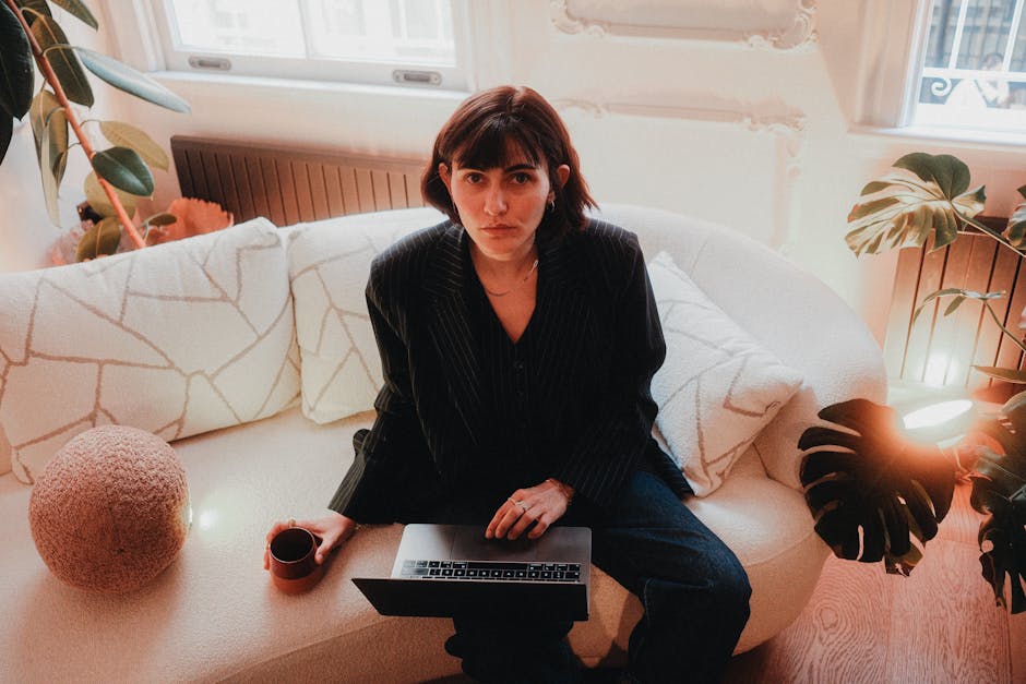 Young woman using a laptop on a couch with warm lighting and indoor plants.