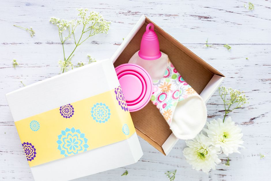 Brightly decorated gift box containing reusable menstrual cup and pads on a rustic background.