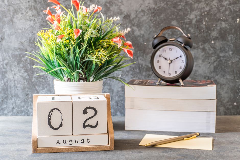 Close-up of a calendar set to August 2nd with a clock, flower vase, and writing pad on a d