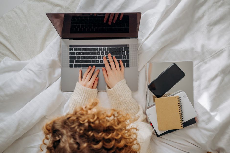 Woman working on a laptop in bed, surrounded by notebooks and smartphone, epitomizing remote work.