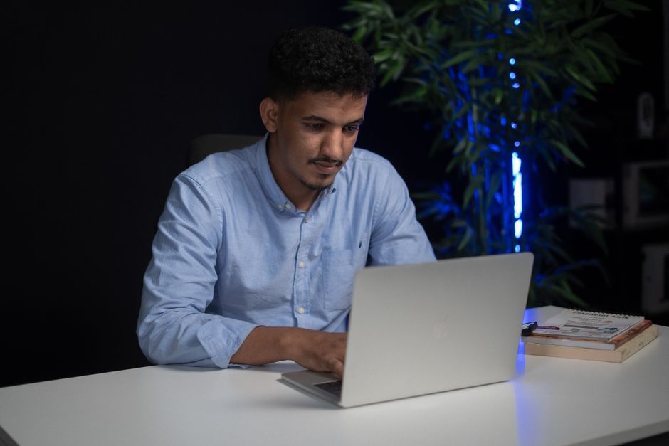 Young man in blue shirt working on a laptop in a dimly lit office with a plant nearby.