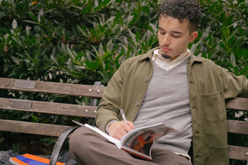 Serious ethnic student male in casual wear writing in workbook while sitting on bench in confident pose in green city park