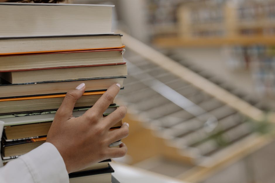 A hand holding a stack of books with bookshelves blurred in the background, depicting knowledge and learning.