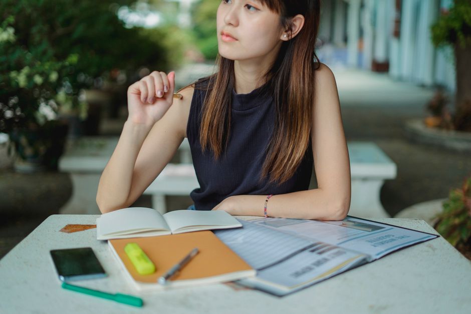Pensive young Asian female thinking while sitting at table and doing homework taking notes in copybook