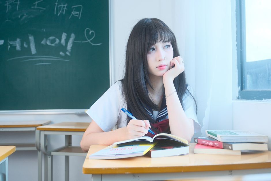 A student in a school uniform sits pensively at a desk in a bright classroom.