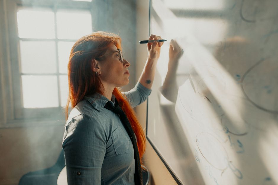 A woman in an office writes on a whiteboard, bathed in natural light streaming through a window.