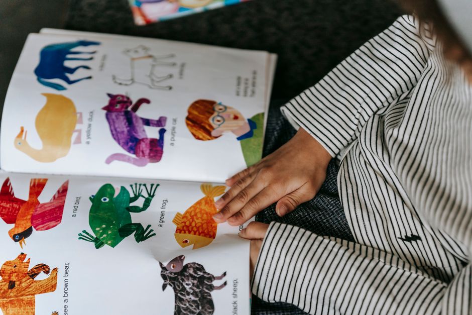 From above of crop anonymous child reading interesting book with colorful pictures of diff