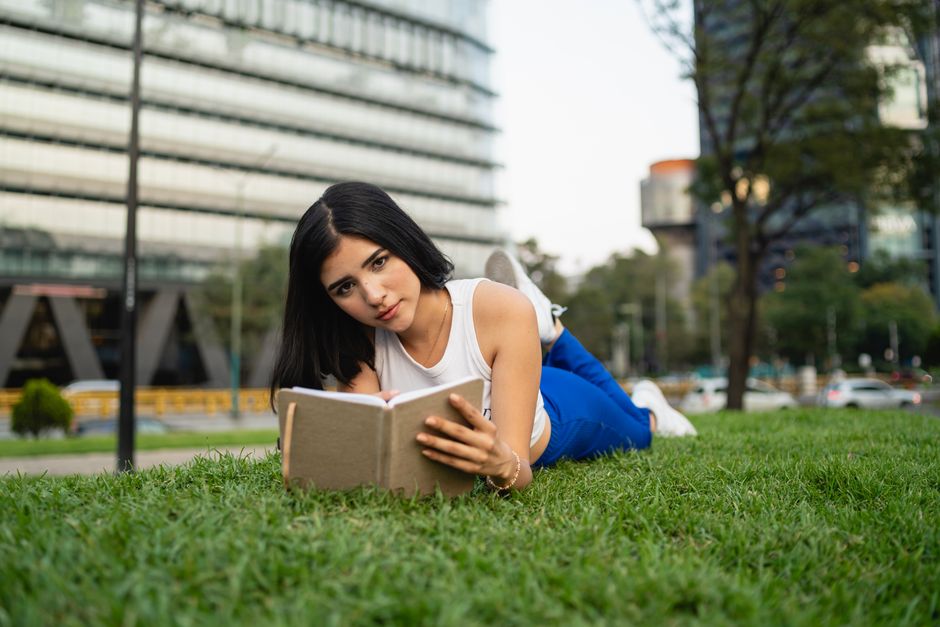 Brunette woman lying on grass reading a notebook in an urban park setting. Fashionable and relaxing scene.