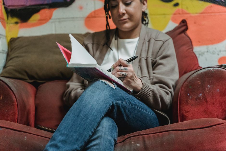 A young woman sitting on a couch and writing in a notebook in a colorful indoor setting.