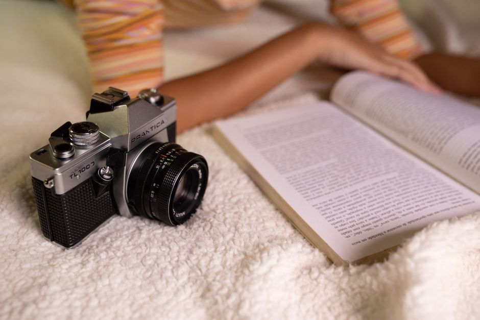 A vintage camera beside an open book on a soft, plush bed indoors.