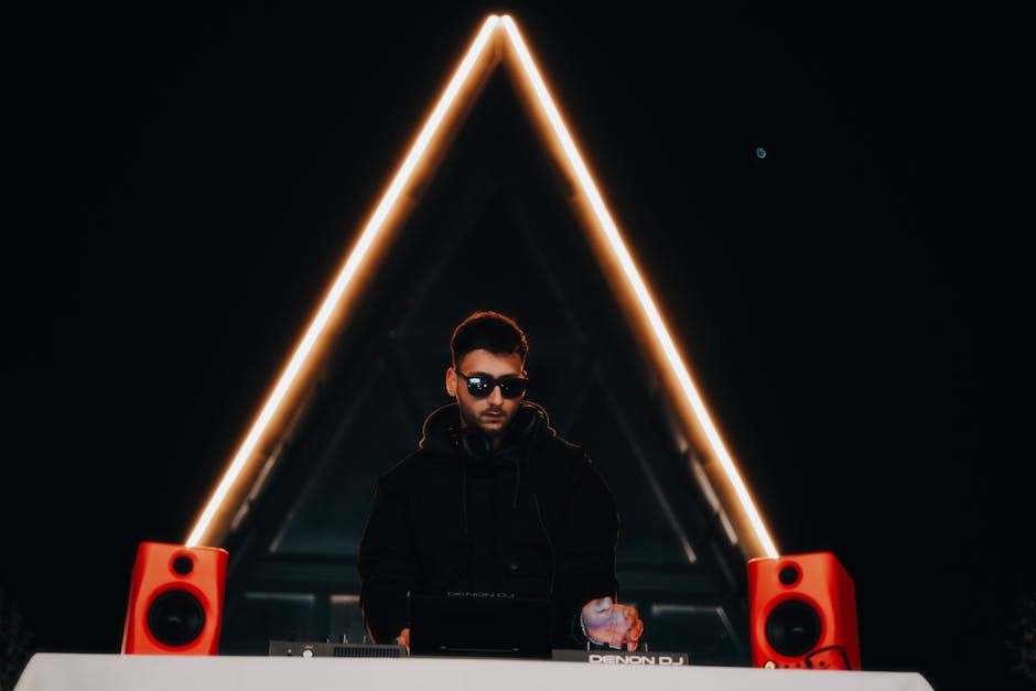 DJ wearing sunglasses plays at a vibrant music event with neon lighting and electronic equipment.