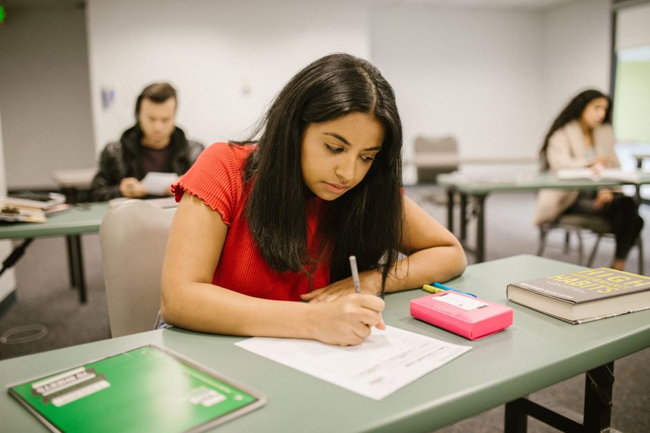 A college student concentrating while writing an exam in a classroom setting, surrounded by peers.