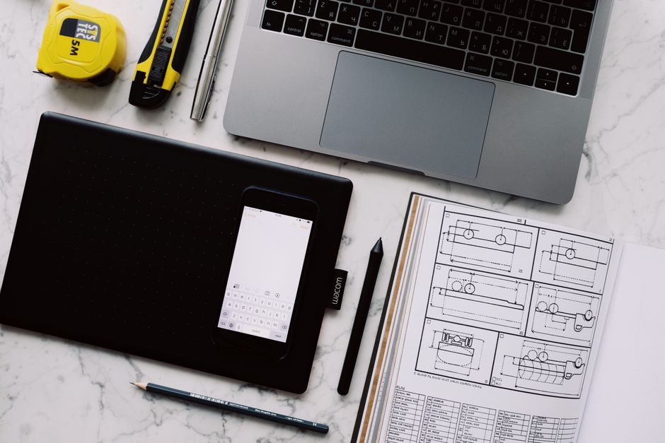 From above view of modern workplace at white marble table with various gadgets and tools and opened notebook with drawings with pen and pencil