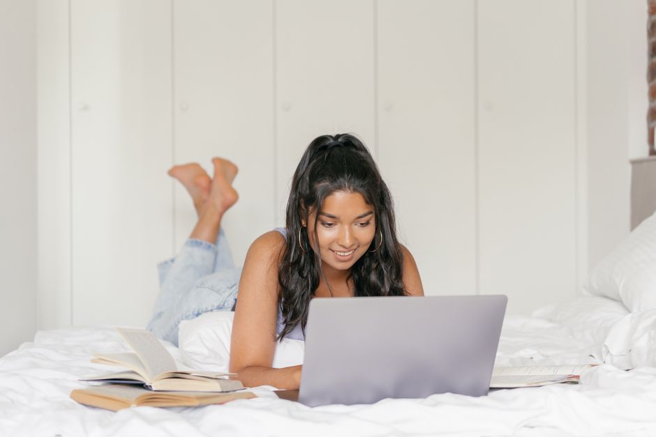 Woman lying on bed with laptop, studying and reading books in a cozy bedroom.
