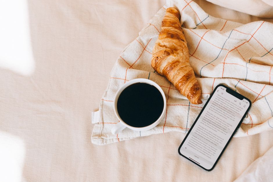 A cozy breakfast in bed scene with black coffee, croissant, and smartphone reading.
