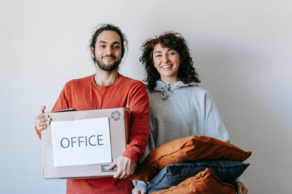 Happy couple preparing for an office move, holding boxes and pillows indoors.