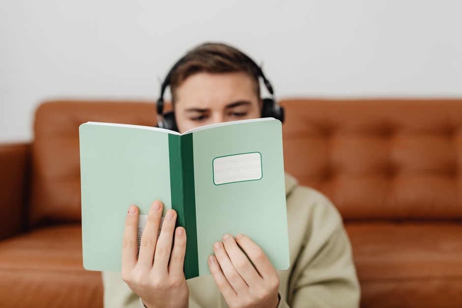 A young man in headphones reading a green notebook on a brown couch, indoors.