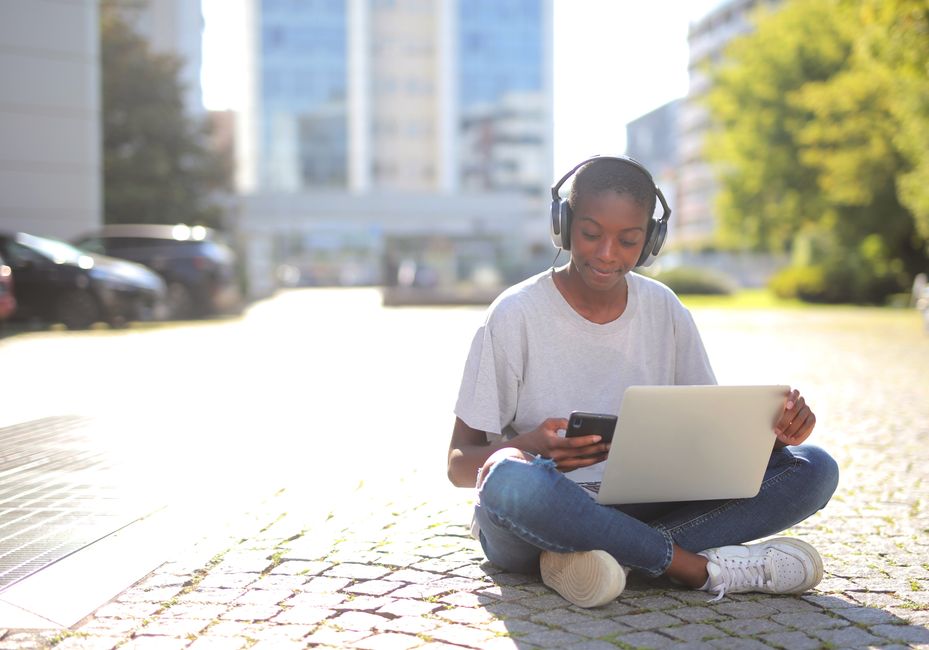Young woman sitting outdoors with headphones, using a laptop and smartphone.