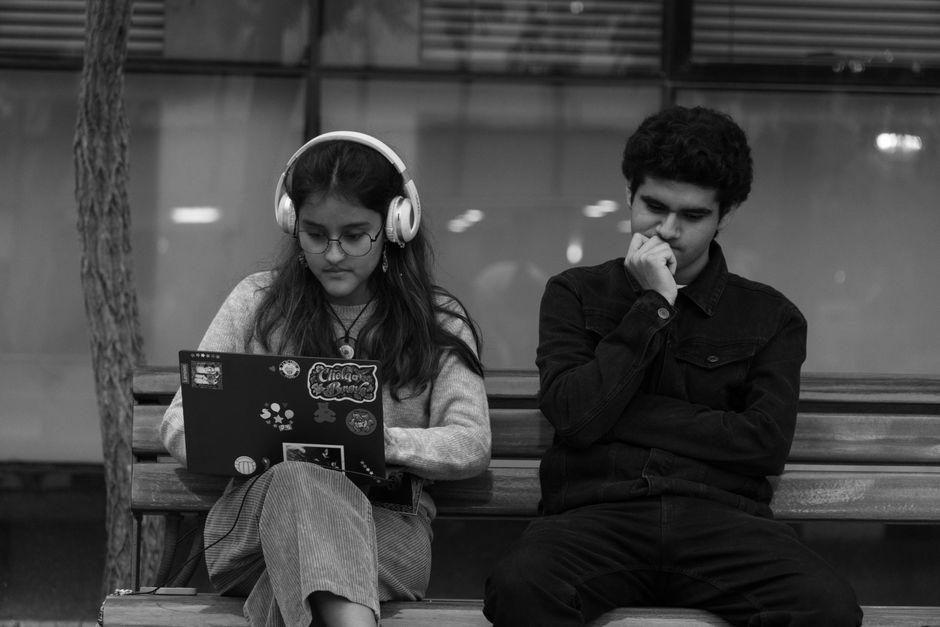 Black and white image of teenagers sitting on a bench, one with headphones and a laptop, the other deep in thought.