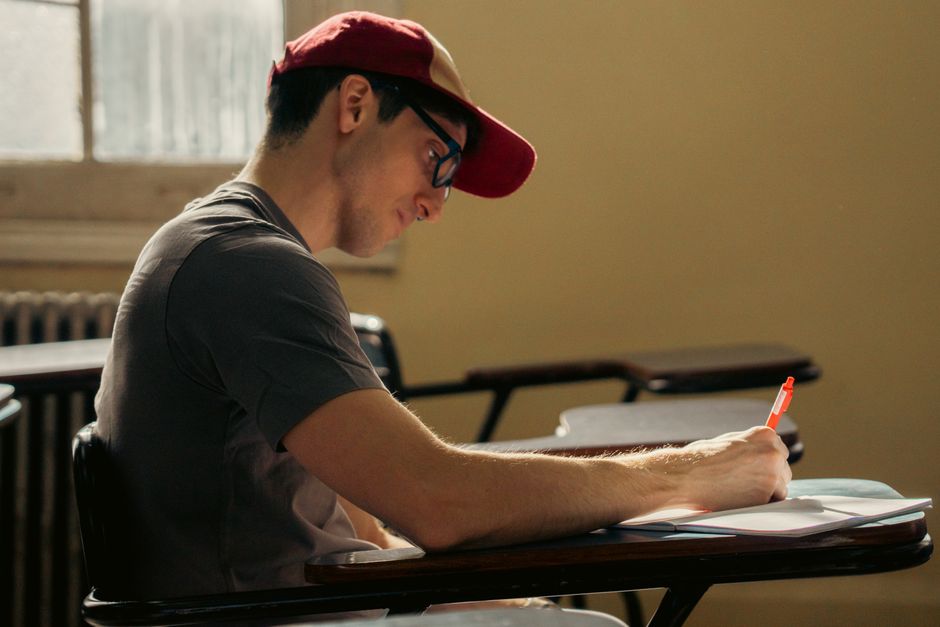 Young male student wearing glasses and cap writing in a classroom.