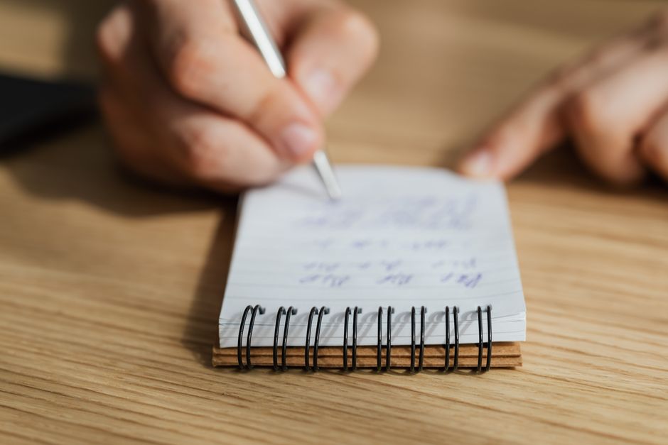 High angle of crop anonymous person taking notes with pen in spiral notebook at wooden table