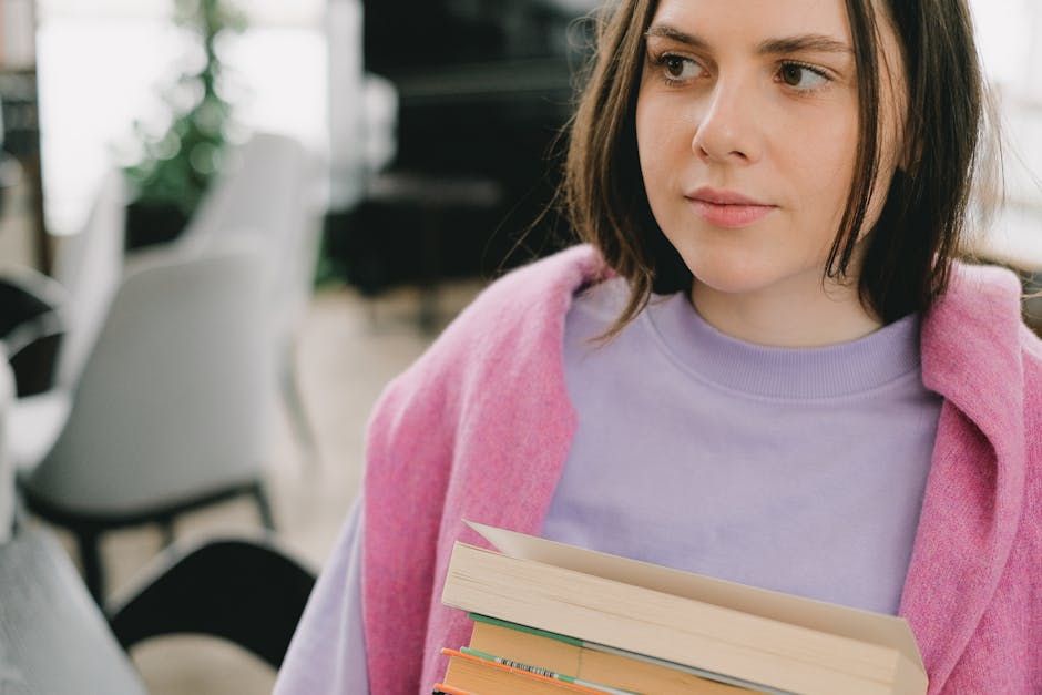 Portrait of a young woman holding books indoors, wearing a pink sweater and looking thoughtful.