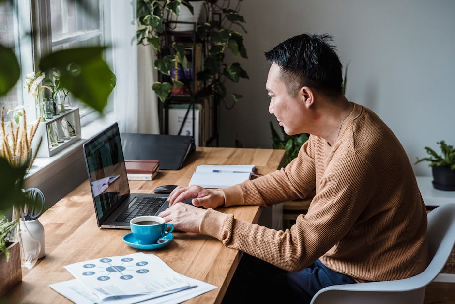 Man in brown sweater working on a laptop with coffee on a wooden desk in a home office setting.